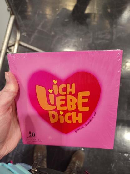 Eine Hand hält ein quadratisches Buch. Rotes Herz auf pinkfarbenem Grund. Darin in gelb: „Ich liebe dich“. Rechts am Herz steht „by M. Lamb, illustrated by Arvi“. Links unten das Logo von „Drei Hasen in der Abendsonne“.