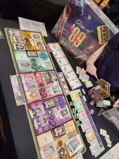 Schräg von oben fotografiertes Spiel „Class of '89“. Hinten steht die Spielepackung, auf der passend zur Zeit neben dem Jahrbuch u.a. eine Musikkassette abgebildet ist. Davor liegt verschiedenes Spielmaterial, u.a. bedruckte Kartenauslagen und Holzmarker, die abgebildeten Personen im Comicstil.