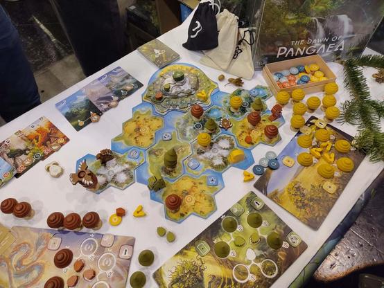 Schräg von oben fotografiertes Spiel „The Dawn of Pangaea“. In der Mitte ein modulares Spielfeld aus Landschsften und Wasser mit Hex-Feldern, auf denen teilweise u.a. Bäume und verschiedenfarbige Waldgeister aus Holz stehen. Am Rand sind Spieletableaus für einzelne Personen zu sehen, auf denen jeweils unterschiedliche Baumkronen und Samen liegen. Dazwischen liegt weiteres Spielmaterial.