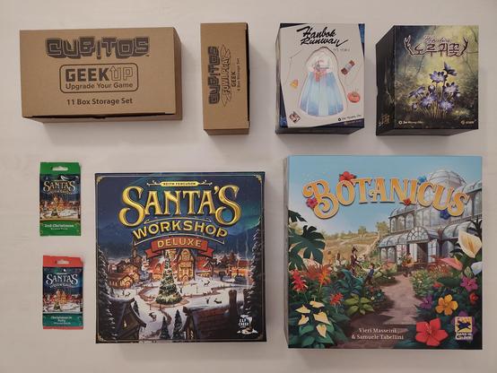 Von oben fotografierte, in zwei Reihen nebeneinander gelegte Spielepackungen. Oben links zwei neutral-braune Pappboxen, bedruckt mit „Cubitos - Geek Up Upgrade Your Game - 11 Box Storage Set“ und „Cubitos Fowl Play - Geek Up - 4 Box Storage Set“. Rechts daneben „Hanbok Runway“, abgebildet sind ein Kleid und Nähmaterial, sowie „Hepatica“, abgebildet sind blaue Blumen auf einer grünen Waldwiese. Unten rechts „Botanicus“ mit einem Gewächshaus, aus der gerade eine Person einen Blumentopf trägt. Davor viele bunte Blumen und ein paar Erwachsene. Links daneben „Santa's Workshop Deluxe“ mit Schriftzug in Gold, darunter ein verschneiter dörflicher Marktplatz mit geschmücktem Weihnachtsbaum in der Mitte. Links am Rand zwei Promo-Packs in Kartengröße für „Santa's Workshop“.