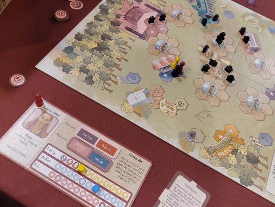 Schräg von oben fotografierter, angeschnittener Spielplan mit einem Festivalgelände, bestehend aus Hex-Feldern, auf denen verschiedene Spielfiguren stehen. . Auf dem Gelände verteilt sind eine Bühne, Getränkestände, Toiletten, Geldautomat und mehr. Vor dem Spielbrett liegt das persönliche Spieltableau mit den besagten drei Punkteskalen sowie der zu Beginn variabel festgelegten Lieblingsmusik.
