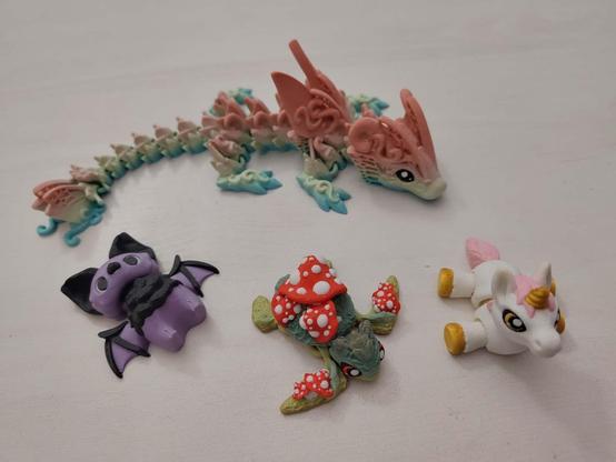 Vier 3D-gedruckte Gliedertiere liegen auf einer weißen Tischplatte, schräg von oben fotografiert. Hinten ein fantasievoll wirkender, länglicher Drache mit Flügeln und Schnörkeln, in Pastellfarben von blau (unten) über grün bis rosa (oben). Davor liegen drei deutlich kleinere Tiere. Links eine Fledermaus in lila und schwarz. In der Mitte eine grpne Schildkröte, bewachsen bon einigen Fliegenpilzen. Rechts ein Einhorn mit ausgestreckten Beinen in weiß mit rosa Mähne und Schweif sowie goldenen Hufen und Horn.
